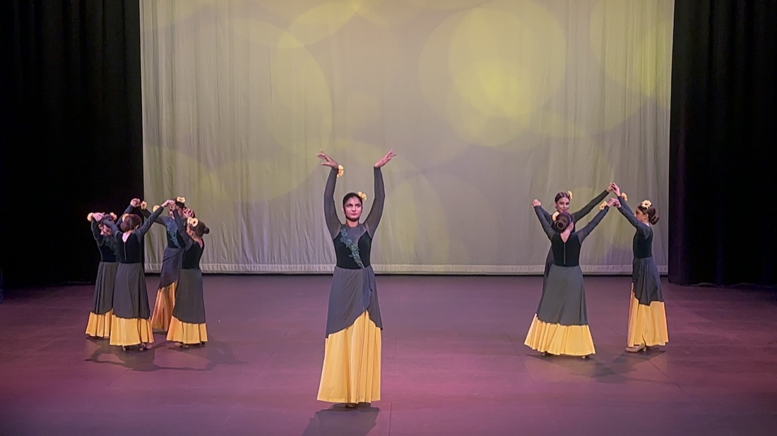 Dancers in gray and gold costumes arranged across the stage, with a central dancer raising both arms.